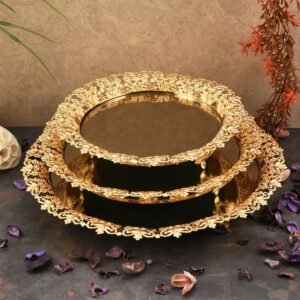 Two-tiered gold display stand with floral designs, dried petals, and plants against a textured wall.