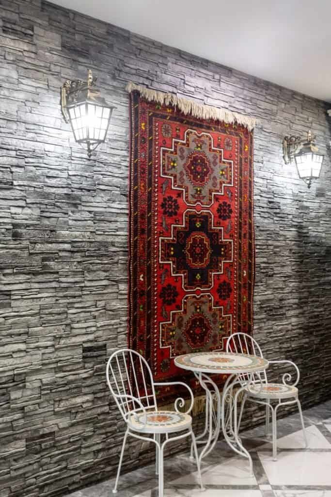 Decorative Scene With Red Tapestry On Stone Wall, Mosaic Table, Two Chairs, And Wall Lamps.